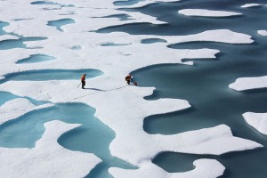 Aerial view of two researchers on a melting ice sheet that is pocketed with large holes, revealing the ocean below. 